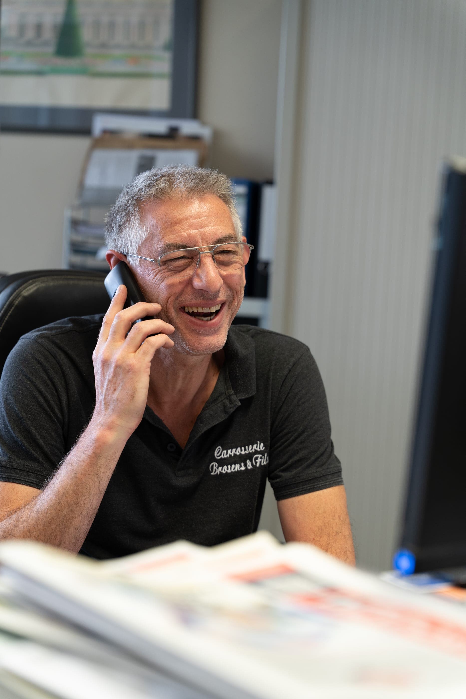 Alain Brosens au téléphone avec un client, souriant, dans le bureau de l’atelier